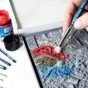 May include: A miniature diorama is being painted with a dry brush. The scene features a detailed, gray, textured base with red, blue, and green accents. A bottle of red paint and a black cap are visible, along with several paint brushes.