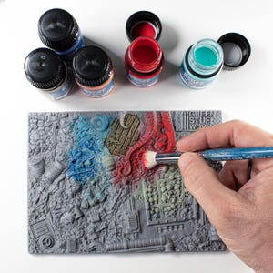 May include: A miniature model being painted with a brush, featuring a detailed, gray, textured surface. Several small bottles of paint in blue, red, and teal are open nearby. The model is partially painted with blue and red hues, showcasing a detailed, textured design.