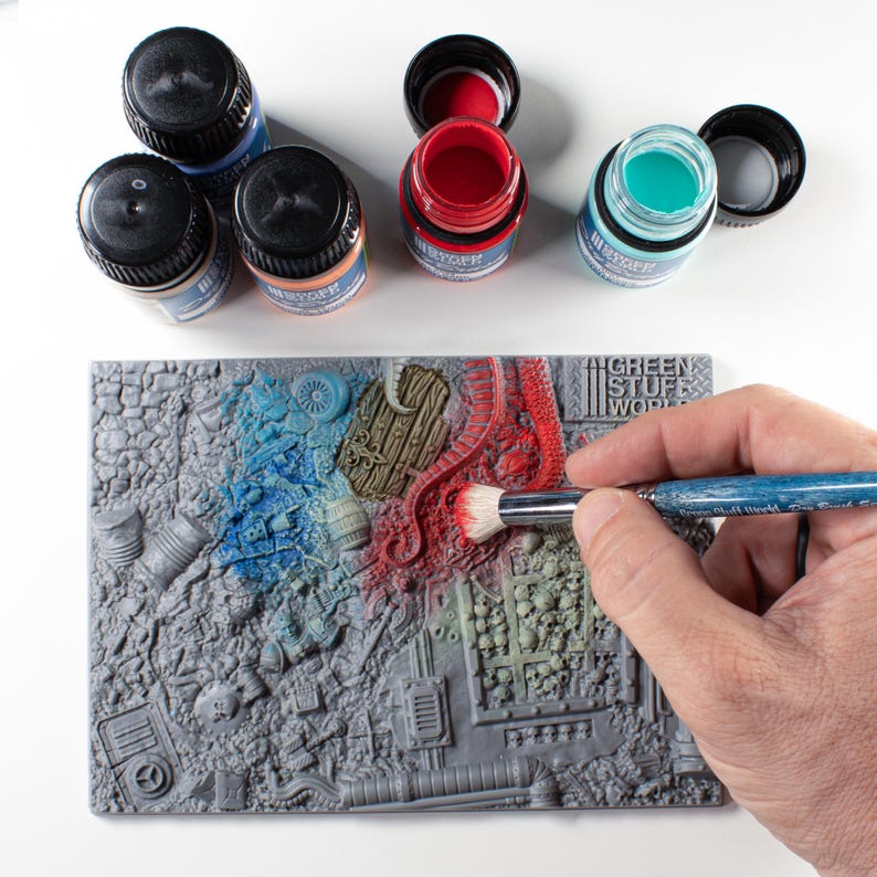 May include: A miniature model being painted with a brush, featuring a detailed, gray, textured surface. Several small bottles of paint in blue, red, and teal are open nearby. The model is partially painted with blue and red hues, showcasing a detailed, textured design.