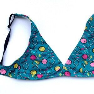 Può includere: Un reggiseno triangolare turchese con un motivo a frutta. Il reggiseno presenta un motivo ripetuto di frutta e foglie rosa, gialle e bianche. Ha spalline regolabili nere e una fascia nera.
