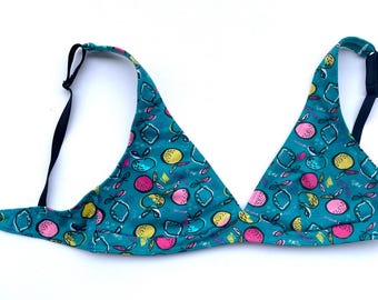May include: A teal triangle bra with a fruit pattern. The bra features a repeating design of pink, yellow, and white fruits and leaves. It has black adjustable straps and a black band.