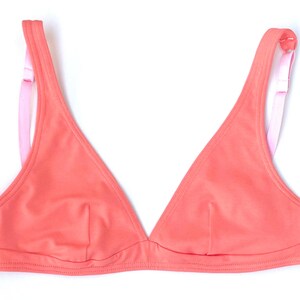 Può includere: Top bikini a triangolo color corallo con spalline rosa. Il top presenta un classico design a triangolo con un sottile dettaglio di cucitura. Le spalline sono di un rosa chiaro a contrasto, aggiungendo un tocco di colore al design generale. Il top del bikini è su sfondo bianco.