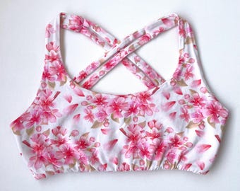 May include: A white sports bra with a pink floral pattern featuring cherry blossoms. The bra has a crisscross back design and is made of a soft, comfortable material. The floral print includes pink flowers and petals.