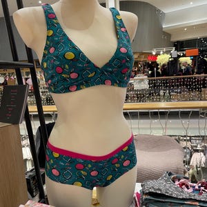 Può includere: Un set di reggiseno e biancheria intima verde acqua su un manichino. Il set presenta un motivo a frutta con accenti rosa, gialli e bianchi. La biancheria intima ha un bordo rosa. Altri set sono impilati su un tavolo.