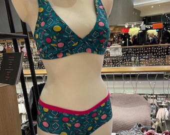 May include: A teal bra and matching underwear set on a mannequin. The set has a fruit pattern with pink, yellow, and white accents. The underwear has a pink trim. Other sets are stacked on a table.