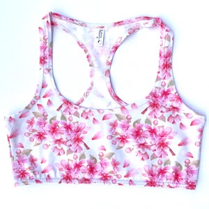 Può includere: Canotta bianca con stampa di fiori di ciliegio rosa. La canotta ha uno stile racerback e una scollatura rotonda.