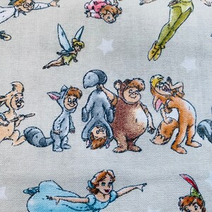 K&ouml;nnte beinhalten: Ein farbenfroher Stoff mit verschiedenen Comicfiguren, darunter Peter Pan, Tinkerbell und Winnie Puuh. Der Stoff ist hellbeige mit einer dezenten Textur.