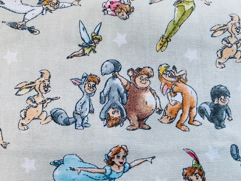 K&ouml;nnte beinhalten: Ein farbenfroher Stoff mit verschiedenen Comicfiguren, darunter Peter Pan, Tinkerbell und Winnie Puuh. Der Stoff ist hellbeige mit einer dezenten Textur.