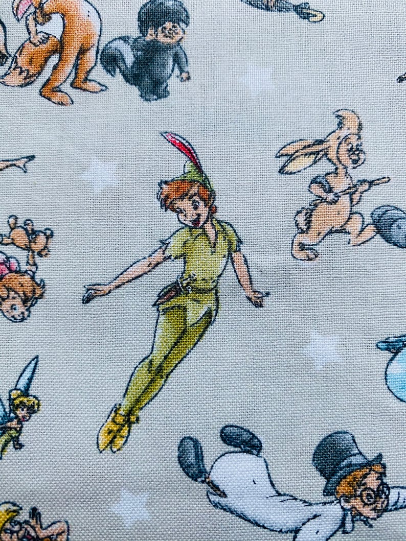 K&ouml;nnte beinhalten: Ein hellgrauer Stoff mit einem sich wiederholenden Muster aus verschiedenen Disney-Charakteren, darunter Peter Pan, Tinkerbell und ein Kaninchen. Die Figuren sind alle im Cartoon-Stil gehalten und fliegen oder stehen auf dem Stoff.