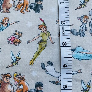 K&ouml;nnte beinhalten: Ein beigefarbener Stoff mit einem sich wiederholenden Muster aus Zeichentrickfiguren aus Peter Pan. Zu den Figuren geh&ouml;ren Peter Pan, Tinkerbell, Captain Hook und die Verlorenen Jungen. Ein wei&szlig;es Ma&szlig;band ist rechts neben dem Stoff platziert und zeigt Ma&szlig;e in Zoll.