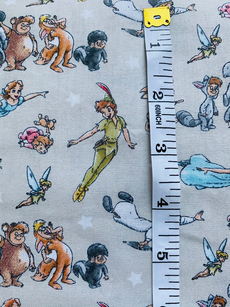 K&ouml;nnte beinhalten: Ein beigefarbener Stoff mit einem sich wiederholenden Muster aus Zeichentrickfiguren aus Peter Pan. Zu den Figuren geh&ouml;ren Peter Pan, Tinkerbell, Captain Hook und die Verlorenen Jungen. Ein wei&szlig;es Ma&szlig;band ist rechts neben dem Stoff platziert und zeigt Ma&szlig;e in Zoll.