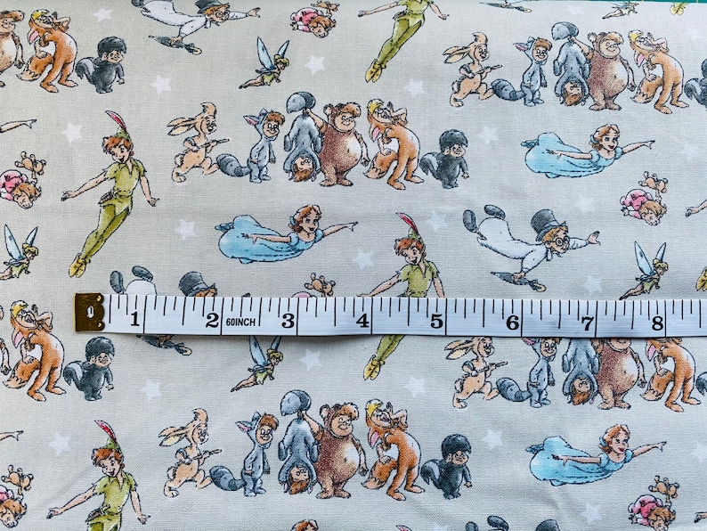 K&ouml;nnte beinhalten: Ein beigefarbener Stoff mit einem sich wiederholenden Muster von Peter Pan und Figuren aus der Geschichte, darunter Tinkerbell, Captain Hook und die Verlorenen Jungen. Der Stoff wird mit einem Lineal gemessen und zeigt 20,3 cm.