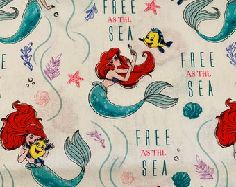 May include: White fabric featuring a repeating pattern of a mermaid with red hair and a teal tail, along with a yellow fish. The phrase "FREE AS THE SEA" is printed in pink. Seaweed and starfish complete the design.