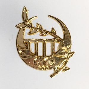 May include: A gold-coloured metal pin featuring a crescent moon design with a stylised lyre inside. The pin has a leafy branch with leaves extending from the top of the crescent moon.