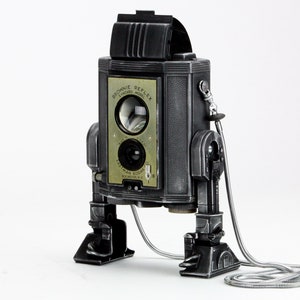 May include: A vintage black and silver Brownie Reflex camera with the text "Brownie Reflex" and "Eastman Kodak" visible. The camera has a unique design with a lens, viewfinder, and a silver cable. The camera is on a white background.