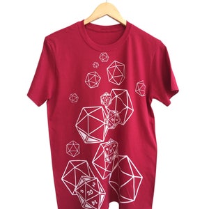 May include: A maroon t-shirt featuring a white graphic design of various polyhedral dice, including D20s, D12s, and D6s. The dice are arranged in a scattered pattern across the front of the shirt. The shirt is hanging on a wooden hanger.