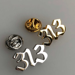 May include: Two silver and gold pins featuring the number 313 in a stylised font. The pins have a safety clasp on the back.