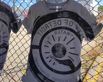 May include: Gray t-shirts with a large black graphic of a Detroit Water Department design. The shirts are hanging on a chain-link fence in front of a blue sky. The graphic includes the text "CITY OF DETROIT WATER DEPARTMENT SEWERAGE MADE IN USA".
