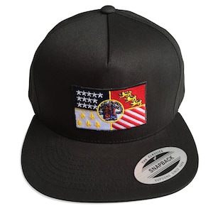 May include: A black and white embroidered flag patch on a black baseball cap. The flag features a red, white, and blue design with a crest in the center. The cap has a black brim and a black strap with a white label that reads "The Classics Snapback Yupoong Since 1974".