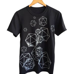 以下が含まれることがあります： 白の幾何学的なサイコロのデザインが施された黒いTシャツ。デザインには、D20、D12、D8など、さまざまな多面体サイコロが含まれています。Tシャツは、白い背景に対して木製のハンガーに掛けられています。