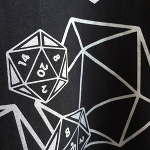 May include: Black fabric with a silver geometric design featuring multiple polyhedral dice. The dice are numbered, with the largest die showing the number 20. The design is a repeating pattern.
