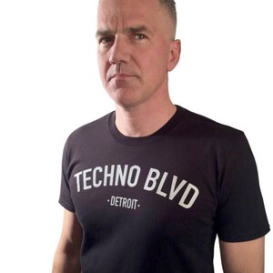K&ouml;nnte beinhalten: Schwarzes T-Shirt mit dem wei&szlig;en Text "TECHNO BLVD - DETROIT" in einem Bogen &uuml;ber der Brust. Die Person auf dem Bild hat kurze, graue Haare und schaut direkt in die Kamera.