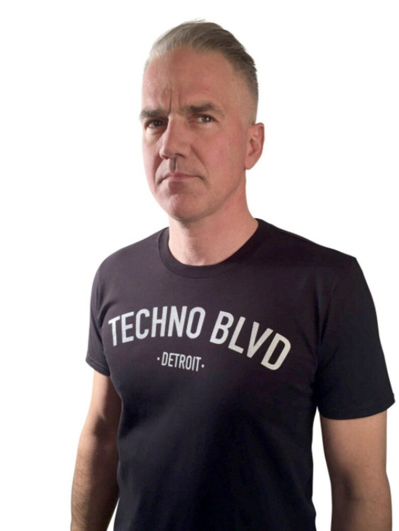 K&ouml;nnte beinhalten: Schwarzes T-Shirt mit dem wei&szlig;en Text "TECHNO BLVD - DETROIT" in einem Bogen &uuml;ber der Brust. Die Person auf dem Bild hat kurze, graue Haare und schaut direkt in die Kamera.