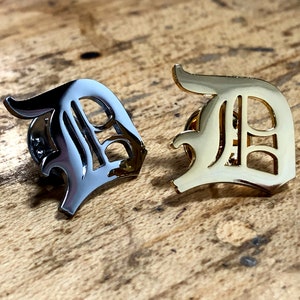 May include: Two metal letter D pins. One is silver and the other is gold. Both are in a gothic font style.