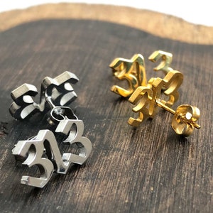 Puede incluir: Un par de pendientes con el número 33 en una fuente gótica. Un par es de plata y el otro es de oro.