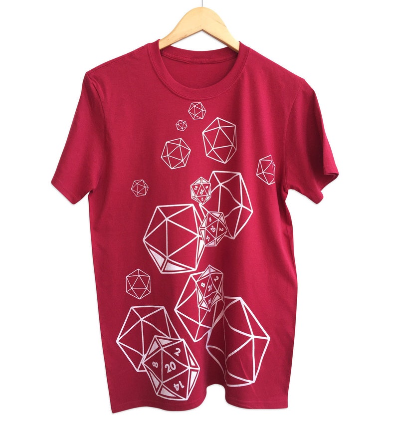 May include: A maroon t-shirt featuring a white graphic design of various polyhedral dice, including D20s, D12s, and D6s. The dice are arranged in a scattered pattern across the front of the shirt. The shirt is hanging on a wooden hanger.