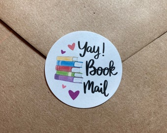 May include: A round sticker with the words "Yay! Book Mail" in black script, with a stack of colorful books and small pink hearts. The sticker is on a brown paper envelope.
