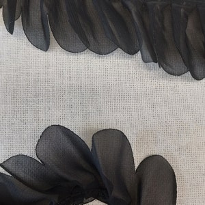 Puede incluir: Adorno de tela negra transparente con bordes festoneados. El adorno está fruncido y plisado, creando un look delicado y femenino.