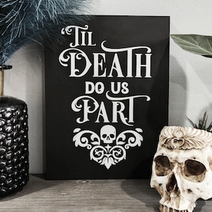 May include: Black rectangular sign with white text that reads "Til Death Do Us Part." The sign features a skull and decorative flourishes. A skull-shaped planter is on the right. The theme is gothic and wedding.