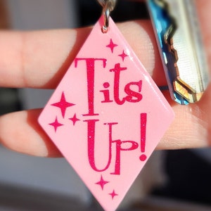 May include: A pink, diamond-shaped keychain with the words "Tits Up!" in glittery pink lettering. The keychain has a silver ring and is held with a key in the background. The design includes star accents.