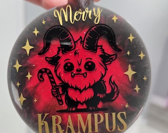 May include: Round Christmas ornament with a red and black background. The ornament features a cartoon Krampus character holding a candy cane. Gold text reads "Merry Krampus" and gold stars are scattered around the edge.