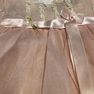 May include: A pale pink dress featuring sheer puffed sleeves, a round neckline trimmed with delicate cream-colored lace, and a satin ribbon waistband tied into a bow on the front. The garment is displayed on a dark blue plastic clothes hanger.