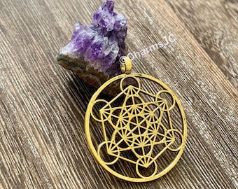 Può includere: Ciondolo in metallo dorato con un design geometrico con dettagli intricati. Il ciondolo ha la forma di un cerchio con una forma geometrica più piccola e dettagliata all'interno. Il ciondolo è su una superficie di legno.