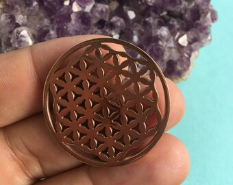 May include: A copper-colored metal pendant featuring a circular design with a geometric pattern known as the Flower of Life. The pendant is resting on a bed of amethyst crystals.