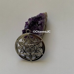 May include: Silver pendant featuring a geometric design with a flower of life pattern. The pendant is on a white surface next to a purple amethyst crystal.