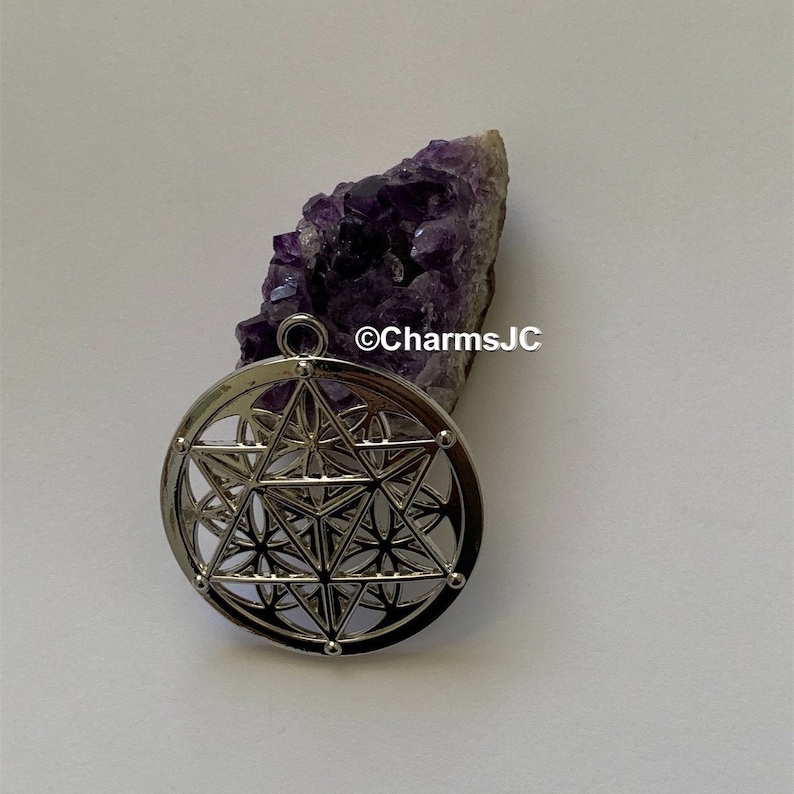May include: Silver pendant featuring a geometric design with a flower of life pattern. The pendant is on a white surface next to a purple amethyst crystal.