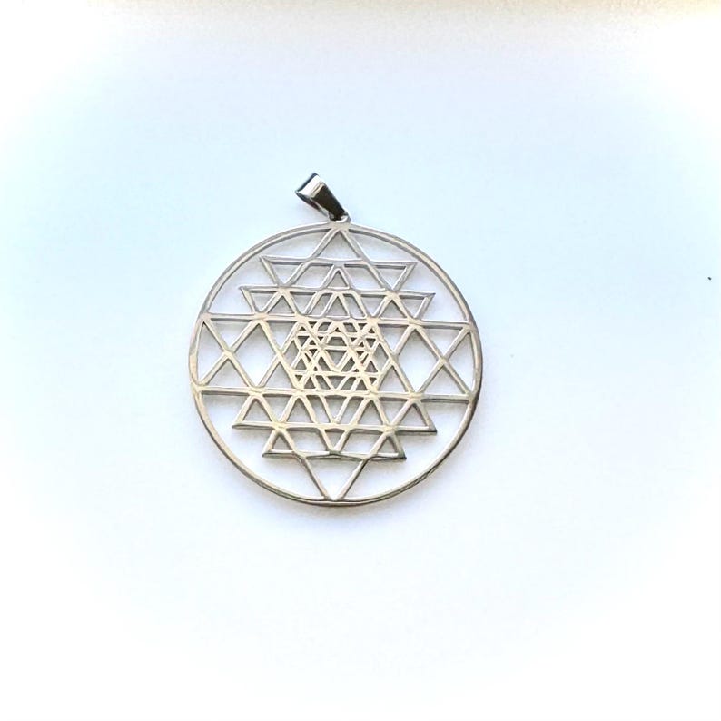 May include: A silver pendant with a geometric Sri Yantra design. The pendant has a circular frame with intricate triangular patterns and a small loop for a chain. The pendant is displayed on a white background.