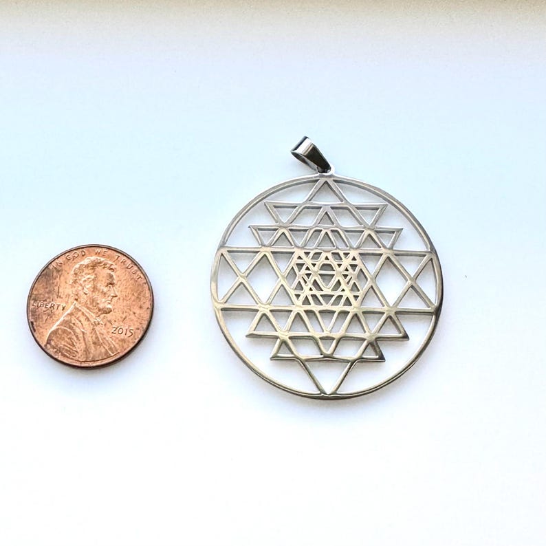 May include: A silver-coloured pendant with a geometric Sri Yantra design, featuring interlocking triangles within a circular frame. A U.S. penny is next to the pendant for size comparison. The pendant has a small loop for a chain.