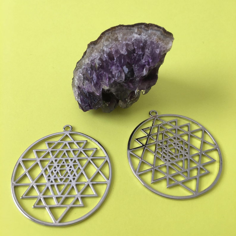 May include: Two silver Sri Yantra pendants, a geometric symbol of sacred geometry, on a yellow background. A purple amethyst crystal is also in the image.