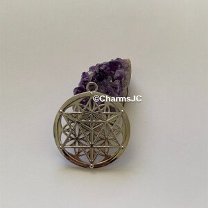 May include: Silver pendant featuring a geometric design with a flower of life pattern. The pendant is on a white background next to a purple amethyst crystal cluster.