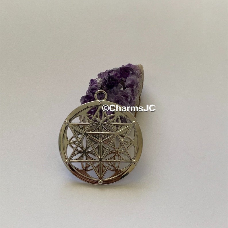May include: Silver pendant featuring a geometric design with a flower of life pattern. The pendant is on a white background next to a purple amethyst crystal cluster.