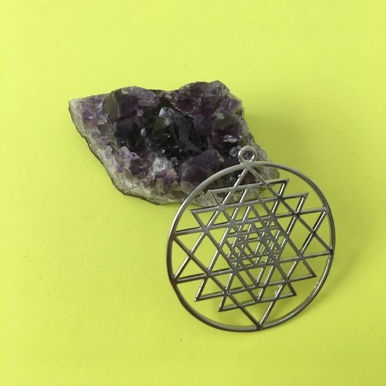May include: A silver Sri Yantra pendant with a geometric design, resting on a purple amethyst crystal cluster against a yellow background.