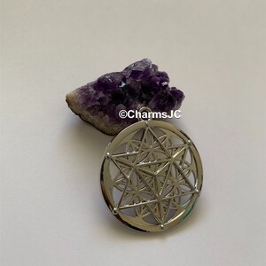 May include: Silver pendant featuring a geometric design with a star in the centre. The pendant is on a white background with a purple amethyst crystal cluster.