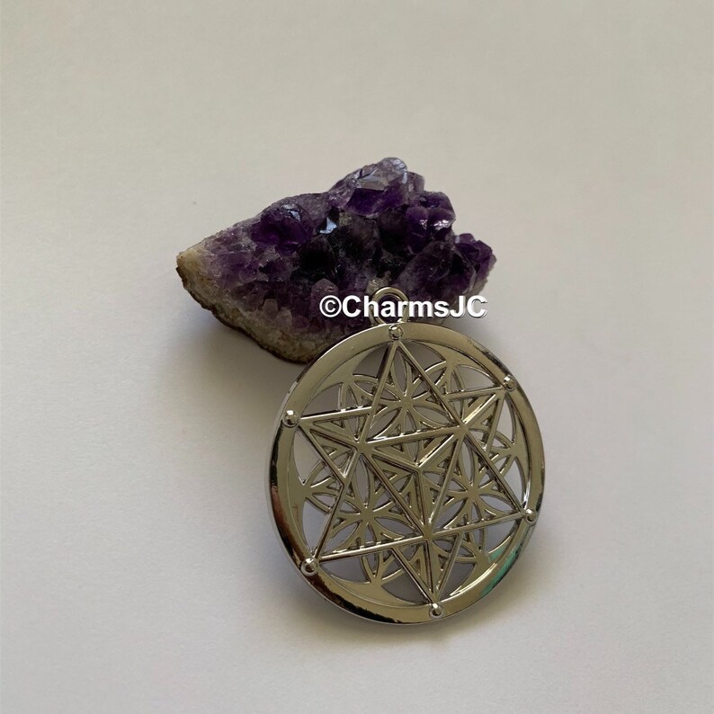 May include: Silver pendant featuring a geometric design with a star in the centre. The pendant is on a white background with a purple amethyst crystal cluster.