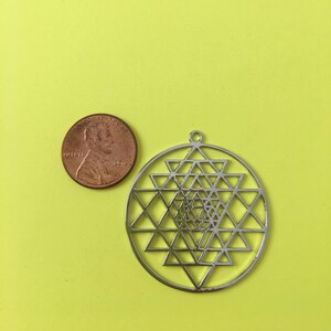 May include: A silver metal pendant featuring a Sri Yantra design. The pendant is approximately the size of a US penny.