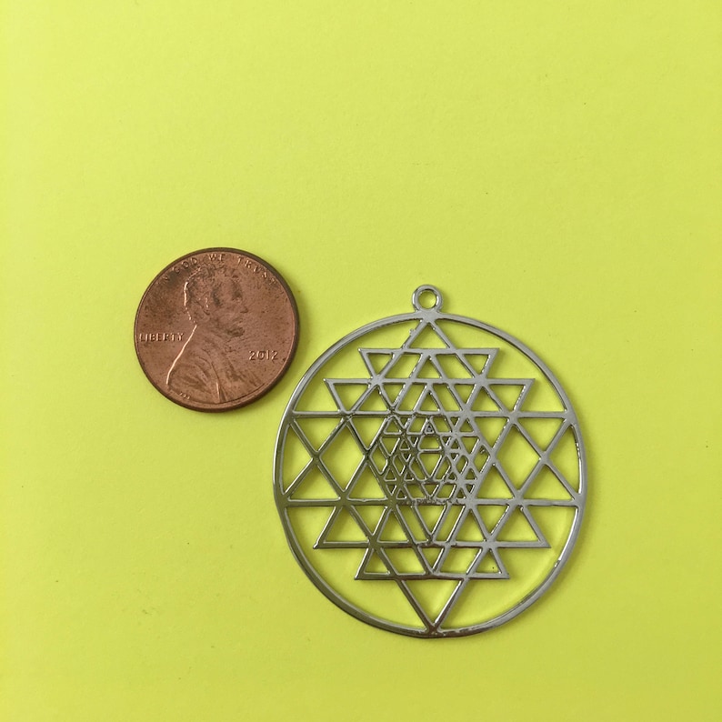 May include: A silver metal pendant featuring a Sri Yantra design. The pendant is approximately the size of a US penny.